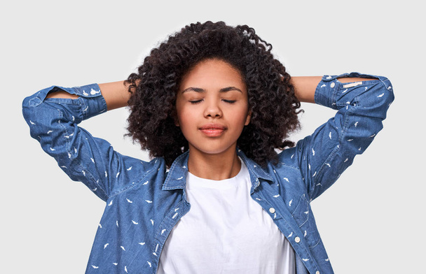 Dreamy dark-skinned young woman dressed in denim shirt and white t-shirt, holding hands on head, feel happy. African American female relaxing with closed eyes posing over white wall - Photo, Image