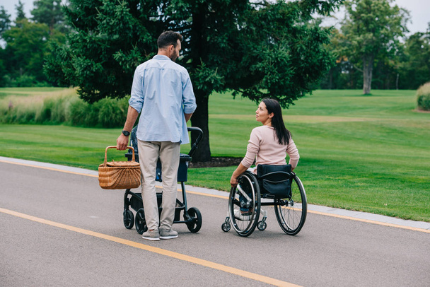 χαρούμενη γυναίκα με αναπηρίες κοιτάζοντας τον άντρα που κουβαλάει καλάθι με άχυρο ενώ περπατάει στο πάρκο με το βρεφικό καροτσάκι μαζί - Φωτογραφία, εικόνα