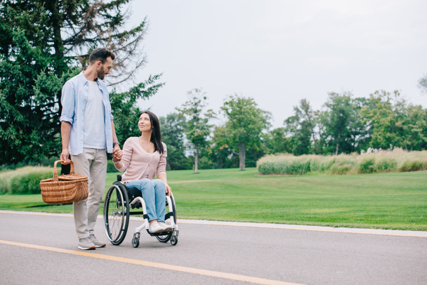 handsome man with straw basket holding hands with disabled girlfriend while walking in park together - Fotoğraf, Görsel