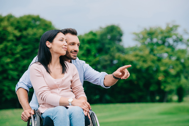 vrolijke man die met de vinger wijst tijdens het wandelen in het park met een gehandicapte vriendin - Foto, afbeelding