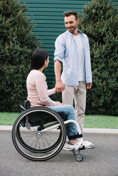 happy man holding hands with disabled girlfriend while walking in park together - Photo, Image