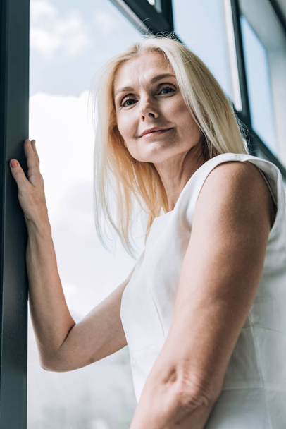 low angle view of elegant blonde mature woman looking at camera near window - Foto, Imagen