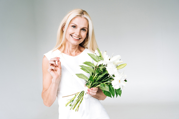 happy blonde mature woman holding white flowers isolated on grey - Foto, Bild