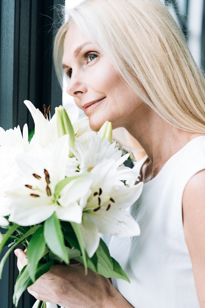 portrait of blonde mature woman with white flowers looking through window - Foto, Imagen