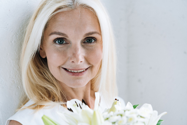 happy blonde mature woman with white flowers near white wall - Zdjęcie, obraz