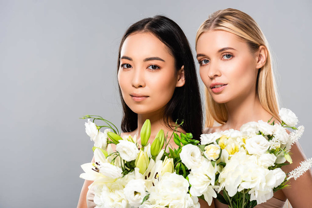 mooie blonde en Aziatische brunette naakte vrouwen met bloemen geïsoleerd op grijs - Foto, afbeelding
