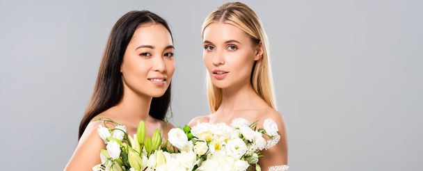 smiling beautiful blonde and asian brunette naked women with flowers isolated on grey, panoramic shot - Fotó, kép