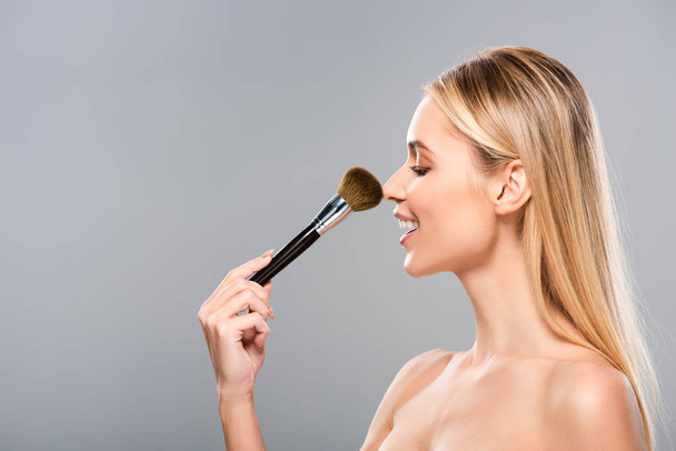 vista lateral de la joven mujer desnuda sonriente con cepillo cosmético aislado en gris
 - Foto, Imagen
