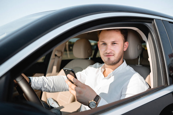 Młody biznesmen uśmiechnięty patrząc na telefon komórkowy podczas jazdy samochodem. - Zdjęcie, obraz