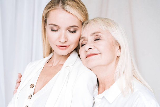 joyful blonde grandmother and granddaughter hugging in total white outfits with closed eyes - Photo, Image