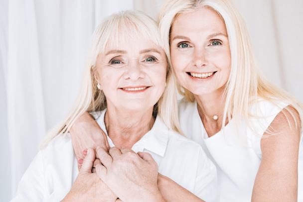 smiling elegant blonde mature daughter and senior mother in total white outfits hugging each other - Фото, зображення