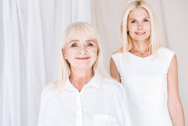 smiling elegant blonde mature daughter and senior mother in total white outfits - 写真・画像