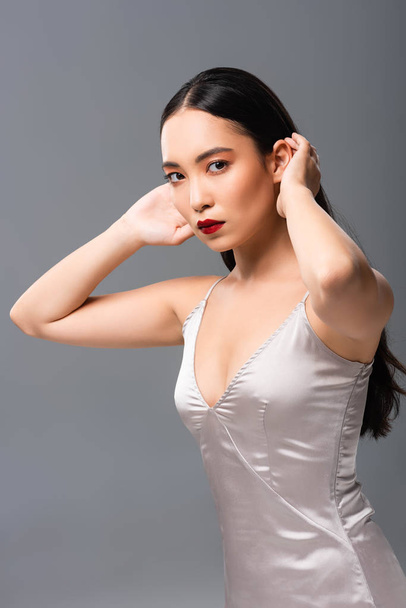 beautiful asian woman in satin dress with red lips touching hair isolated on grey - Photo, Image