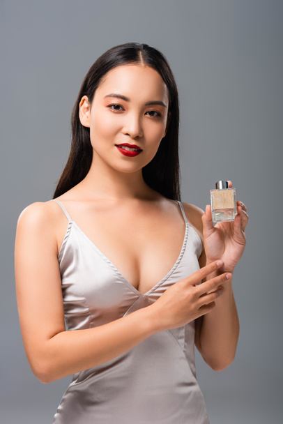 beautiful asian woman in elegant dress with red lips holding perfume isolated on grey - Photo, Image