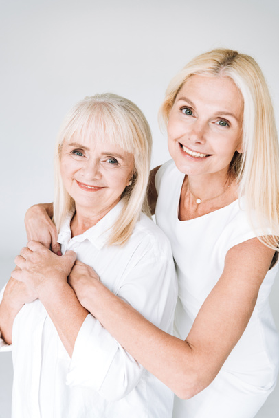 elegant blonde mature daughter and senior mother in total white outfits embracing isolated on grey - 写真・画像