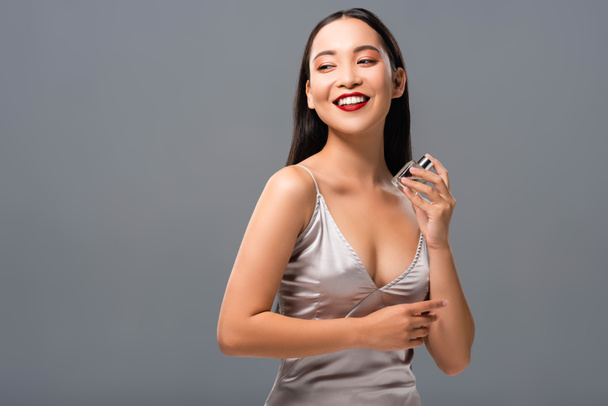 beautiful smiling asian woman with red lips holding perfume isolated on grey - Photo, Image