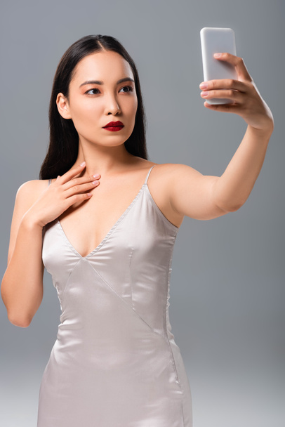 kaunis aasialainen nainen satiini mekko punaiset huulet ottaen selfie eristetty harmaa
 - Valokuva, kuva