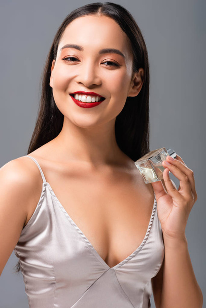 krásná usměvavá asijská žena v elegantních šatech s červenými rty, které drží parfémy izolované na šedé - Fotografie, Obrázek
