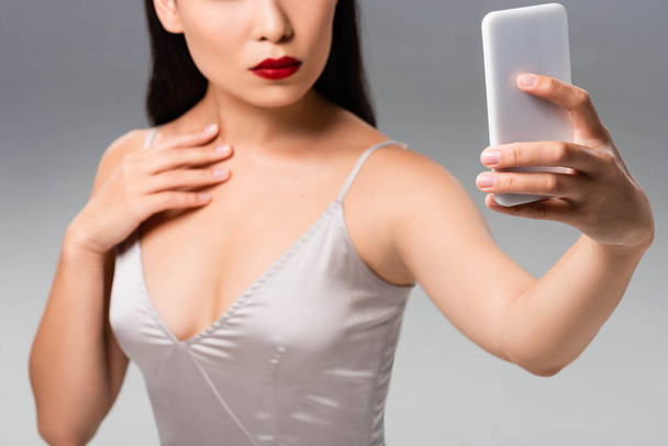 cropped view of beautiful woman in satin dress with red lips taking selfie isolated on grey - Fotó, kép