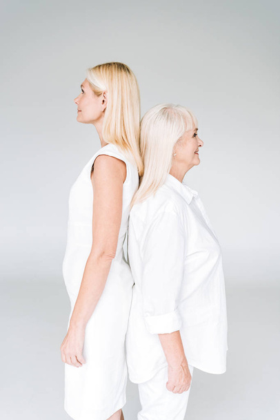 blonde mature daughter and senior mother in total white outfits standing back to back isolated on grey - Foto, afbeelding