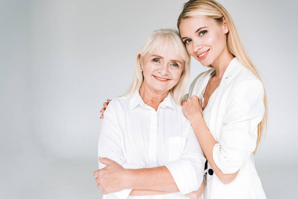 happy blonde grandmother and granddaughter together in total white outfits embracing isolated on grey - Photo, Image