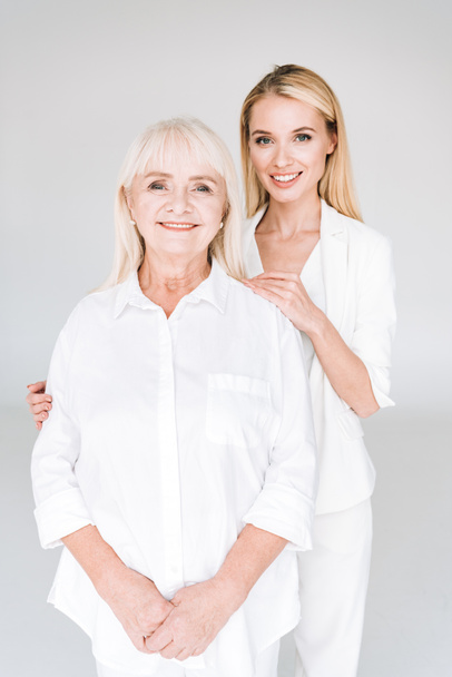 smiling blonde grandmother and granddaughter together in total white outfits embracing isolated on grey - Photo, Image
