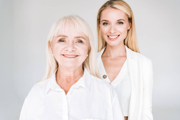 smiling blonde grandmother and granddaughter together in total white outfits isolated on grey - Photo, Image
