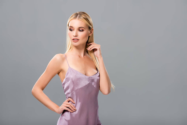 Elegancka Blondynka w fioletowej satynowej sukni patrząc w dół z ręką na biodrze izolowane na szarym - Zdjęcie, obraz