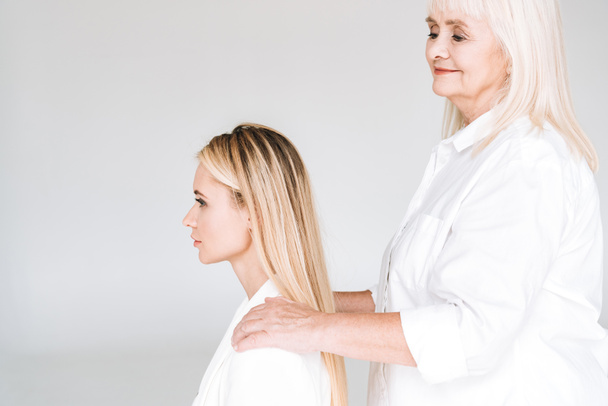 side view of blonde grandmother putting hands on granddaughter shoulders isolated on grey - Fotó, kép