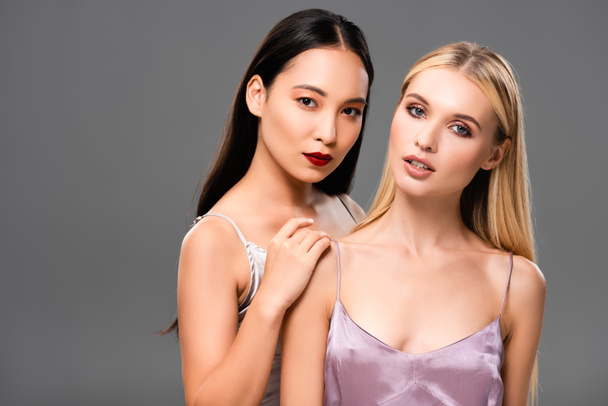 beautiful european blonde and asian brunette women in satin dresses isolated on grey - Photo, Image