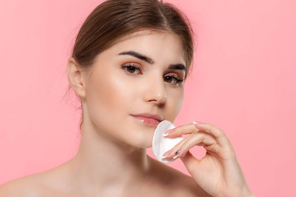 Beautiful model applyes cosmetic product to skin with white cotton sponge. Model looks at the camera isolated over pink background. - Photo, Image