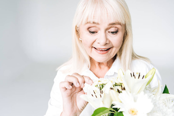 exited blonde senior woman in white clothes with flowers isolated on grey - Photo, Image