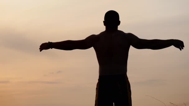 Pohled z pozadí siluety atletického muže s obnaženými trupem ukazuje jeho svaly a biceps na oblohu při západu slunce. Sport, zdraví a fitness - Záběry, video