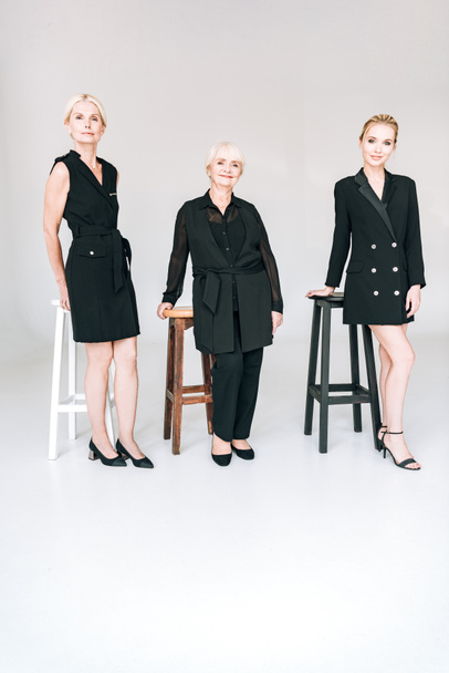vista a tutta lunghezza di donne bionde alla moda di tre generazioni in abiti neri totali vicino a sedie isolate sul grigio
 - Foto, immagini