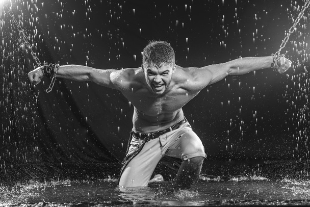 Strong man in chains posing under the rain, aqua studio - 写真・画像