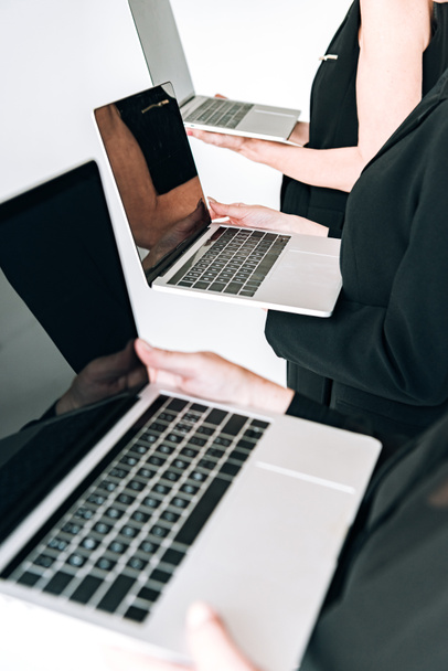 vista recortada de mulheres de negócios de três gerações em roupas pretas totais segurando laptops com telas em branco isoladas em cinza
 - Foto, Imagem