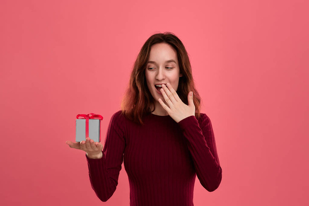 Excited pretty brunette girl holding present box with a ribbon standing isolated on a dark pink background and smiling at the camera. Celebrating special event. - Fotografie, Obrázek