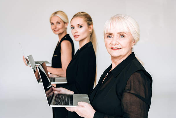 Widok z boku modne trzy pokolenia blond bizneswomen w sumie Czarne stroje gospodarstwa laptopy izolowane na szarym - Zdjęcie, obraz