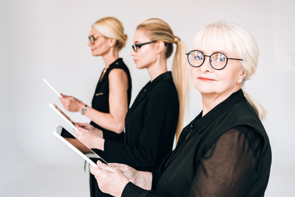 mujeres de negocios rubias de moda de tres generaciones en trajes negros totales y gafas que sostienen tabletas digitales aisladas en gris
 - Foto, Imagen
