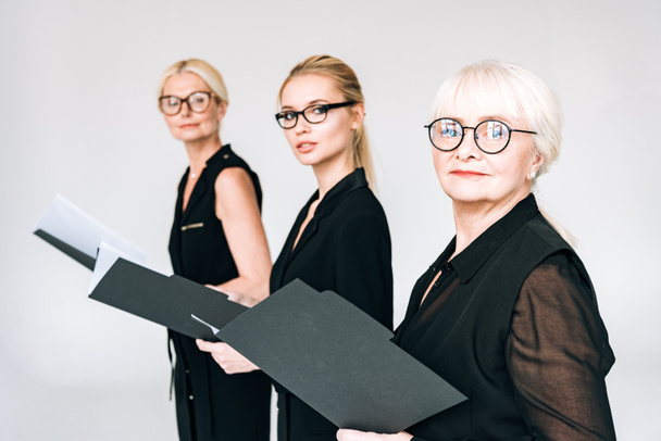 модные женщины-бизнесмены трех поколений в очках с папками, изолированными на сером
 - Фото, изображение