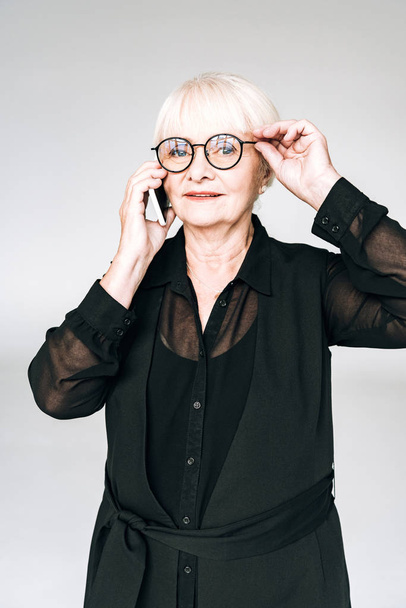 senior businesswoman in black outfit and glasses talking on smartphone isolated on grey - Photo, Image