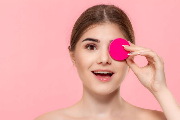 Krásný model tleskne kosmetickým přípravkem na kůži s růžovou bavlněnou houbou. Model vyhledává fotoaparát izolovaný na růžovém pozadí. - Fotografie, Obrázek