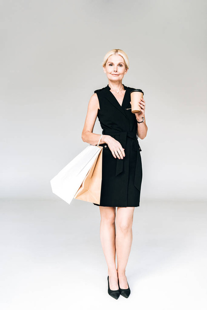 teljes hosszúságú tekintettel az elegáns szőke nő, fekete ruhában a bevásárló táskák és kávé menni izolált szürke - Fotó, kép