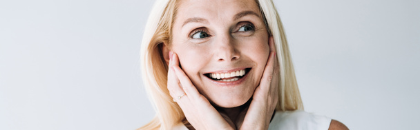 panoramatický záběr šťastné blonďaté ženy s rukama na tváři, která se dívá jinam na šedou - Fotografie, Obrázek