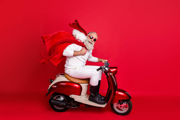 Foto lateral de perfil de longitud completa del divertido pensionista que lleva el bolso con regalos sentado en la motocicleta que se apresura a usar botas de pantalones de suéter blanco aisladas sobre fondo rojo
 - Foto, imagen