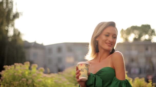 Close up of girl with blond hair laughing looking at camera. Cityscape with trees. With a paper cup of coffee in hand. - Materiał filmowy, wideo