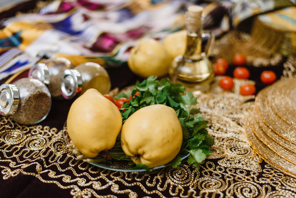 Owoce Quince na stole ozdobione orientalnymi tkaninami z ozdobą i złotymi akcentami. Koncepcja kuchni orientalnej. Owoce Quince, pomidory i zioła są na stole. - Zdjęcie, obraz