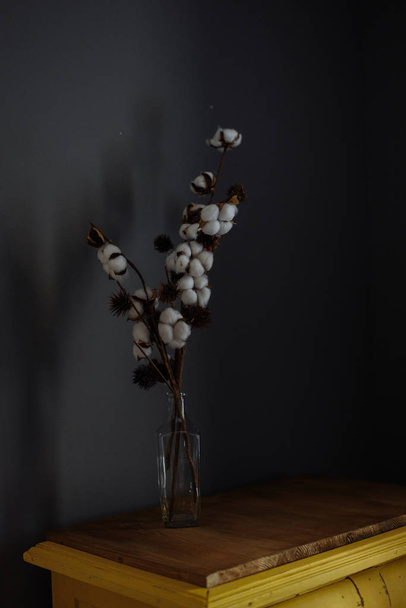 decorative branch with cotton stands in a transparent vase on a wooden chest. Simple and dark room interior - Foto, Bild