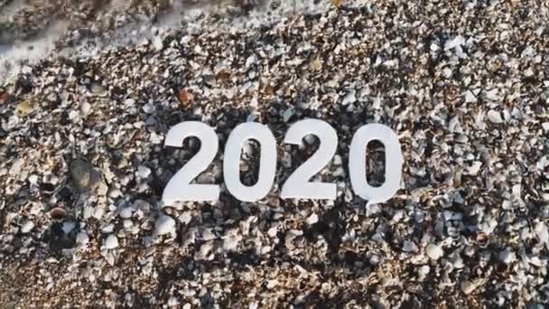 2020 yılı kumdaki sayılar, deniz kıyısındaki taş ve kabukların arasında bir deniz dalgası tarafından yıkanır. Yeni Yıl 2020 kavramı geliyor - bir kum plajda rakamlar. Yeni yıl tatili. Close. Üstte görüntü.  - Video, Çekim