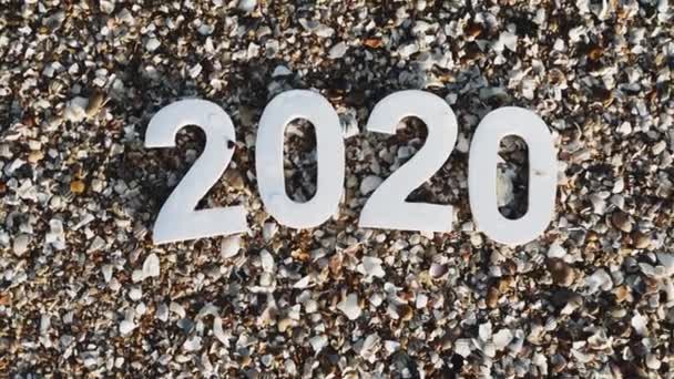 2020 yılı kumdaki sayılar, deniz kıyısındaki taş ve kabukların arasında bir deniz dalgası tarafından yıkanır. Yeni Yıl 2020 kavramı geliyor - bir kum plajda rakamlar. Yeni yıl tatili. Close. Üstte görüntü.  - Video, Çekim